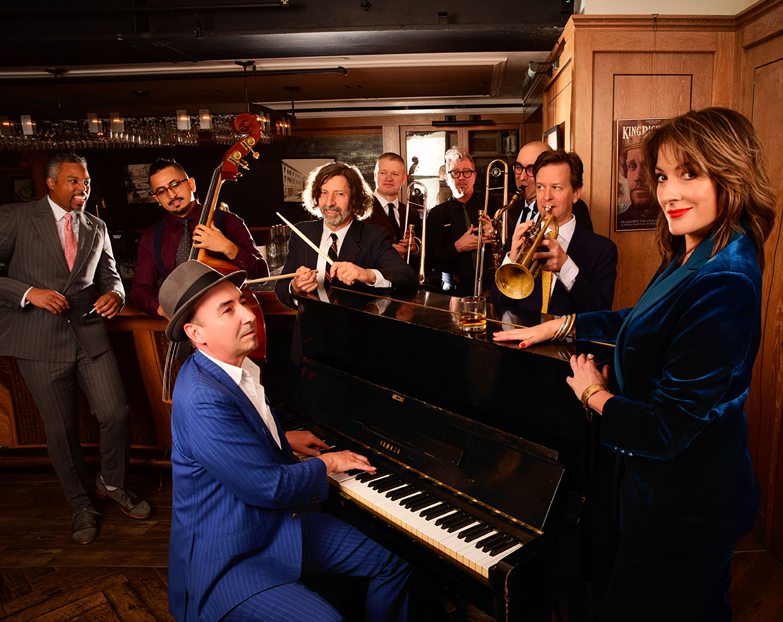 An ensemble photo of the Hot Sardines, a jazz group of eight men and one woman gathered around a piano with their instruments.