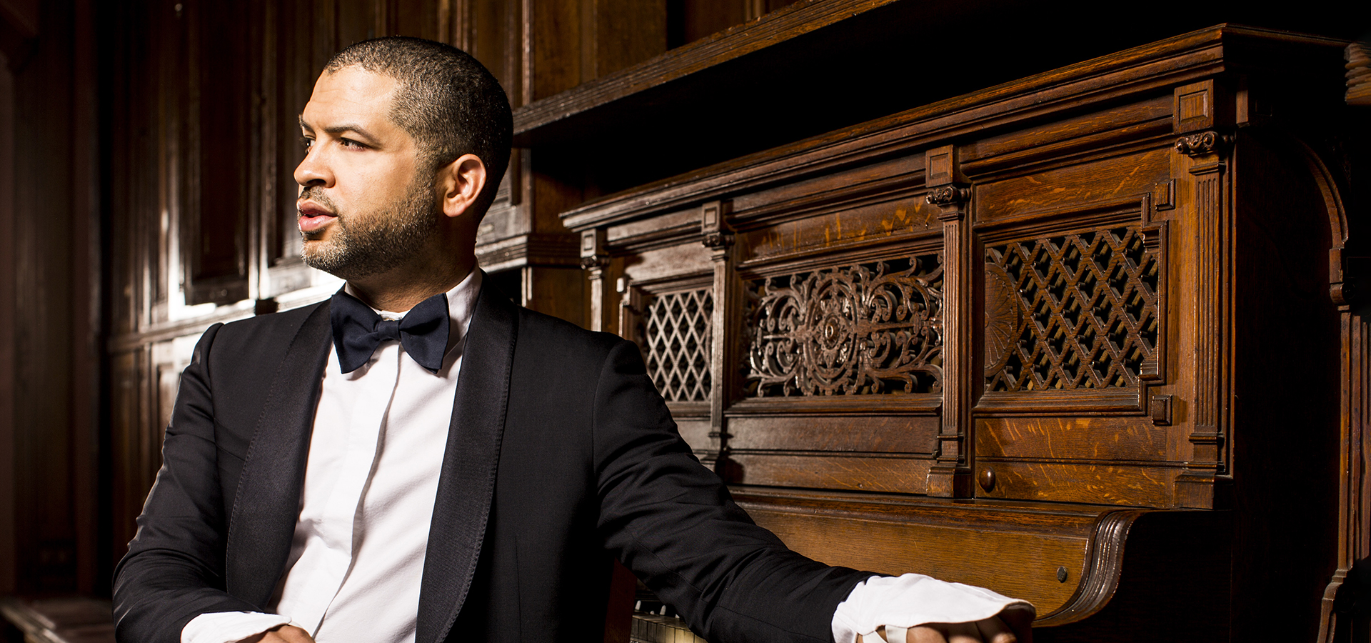 Jazz pianist Jason Moran, a black man in a black suit and bow tie, looks to the side and leans agaist an upright piano.