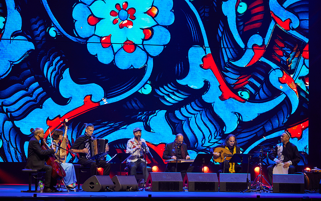 An ensemble photo of instrumentalists of Aga Khan Master Musicians in concert on stage in front of a blue and red backdrop.