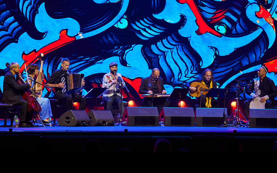 An ensemble photo of instrumentalists of Aga Khan Master Musicians in concert on stage in front of a blue and red backdrop.