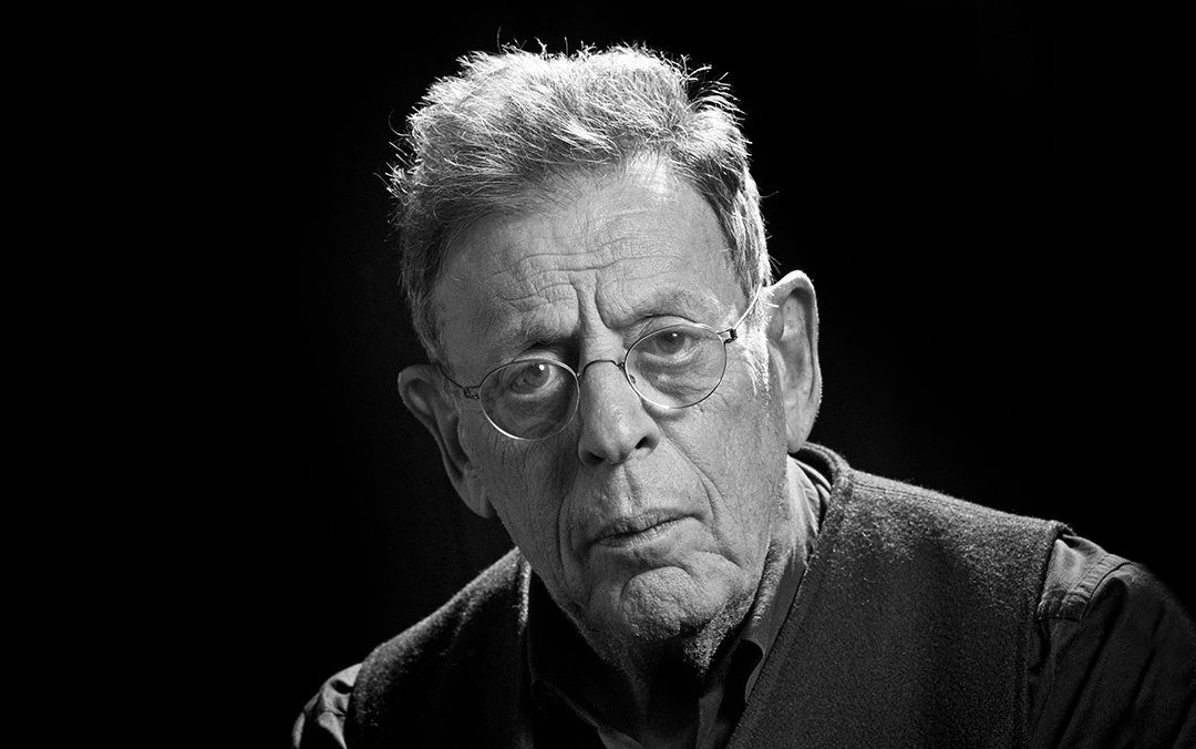 A black and white portrait of Philip Glass, an older man in a dark shirt and vest wearing glasses looking directly into the camera.