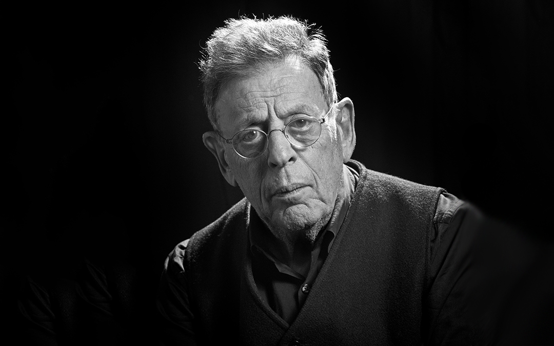 A black and white portrait of Philip Glass, an older man in a dark shirt and vest wearing glasses looking directly into the camera.