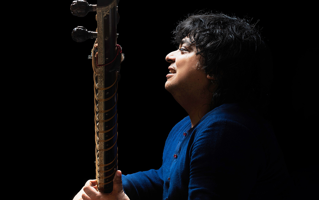 Sitarist Niladri Kumaar, a south Asian man dressed in blue sitting sideways and holding his instrument.