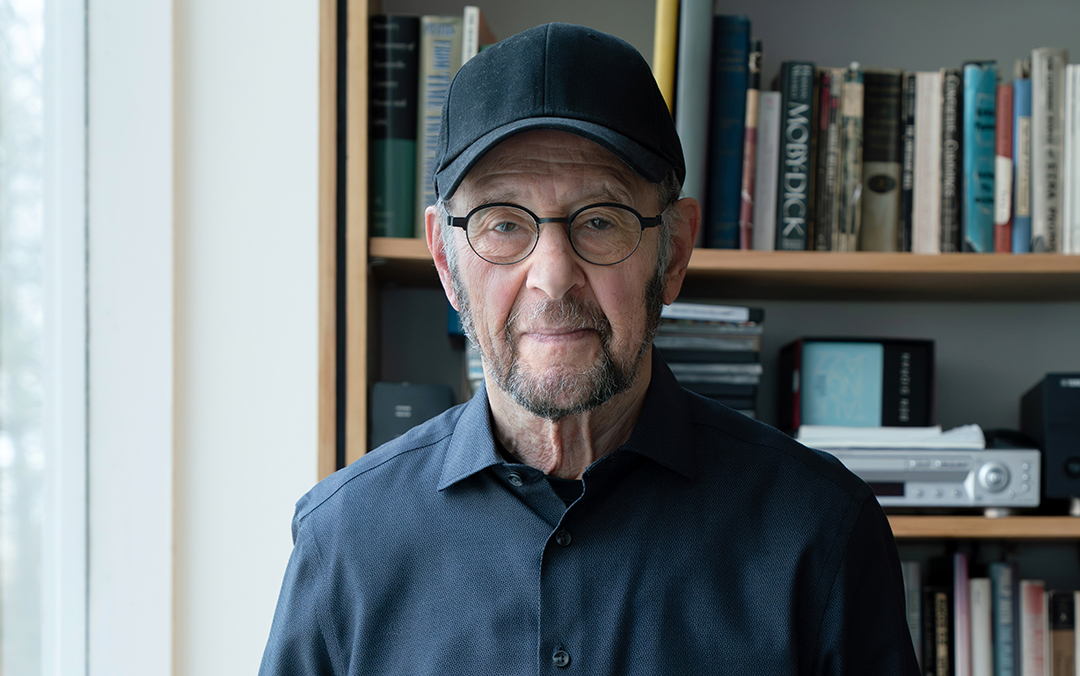 Composer Steve Reich, an older man with glasses and a baseball hat stands in front of a bookcase filled with books.