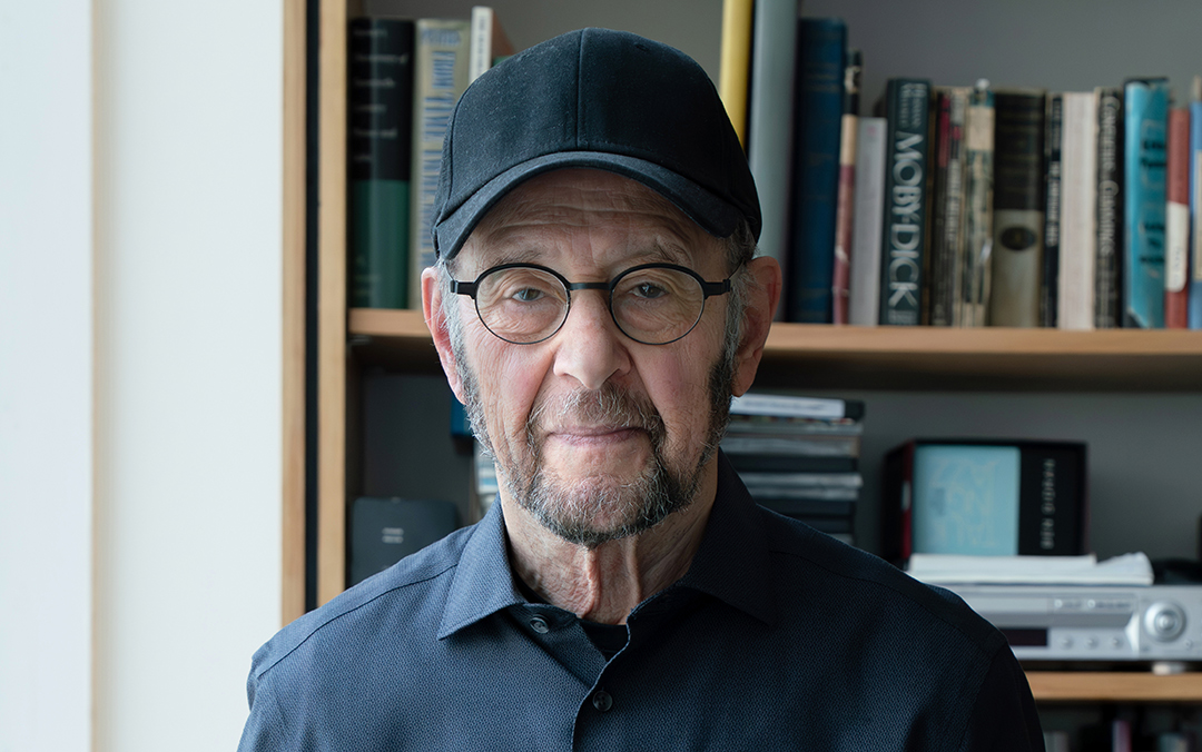 Composer Steve Reich, an older man with glasses and a baseball hat stands in front of a bookcase filled with books.