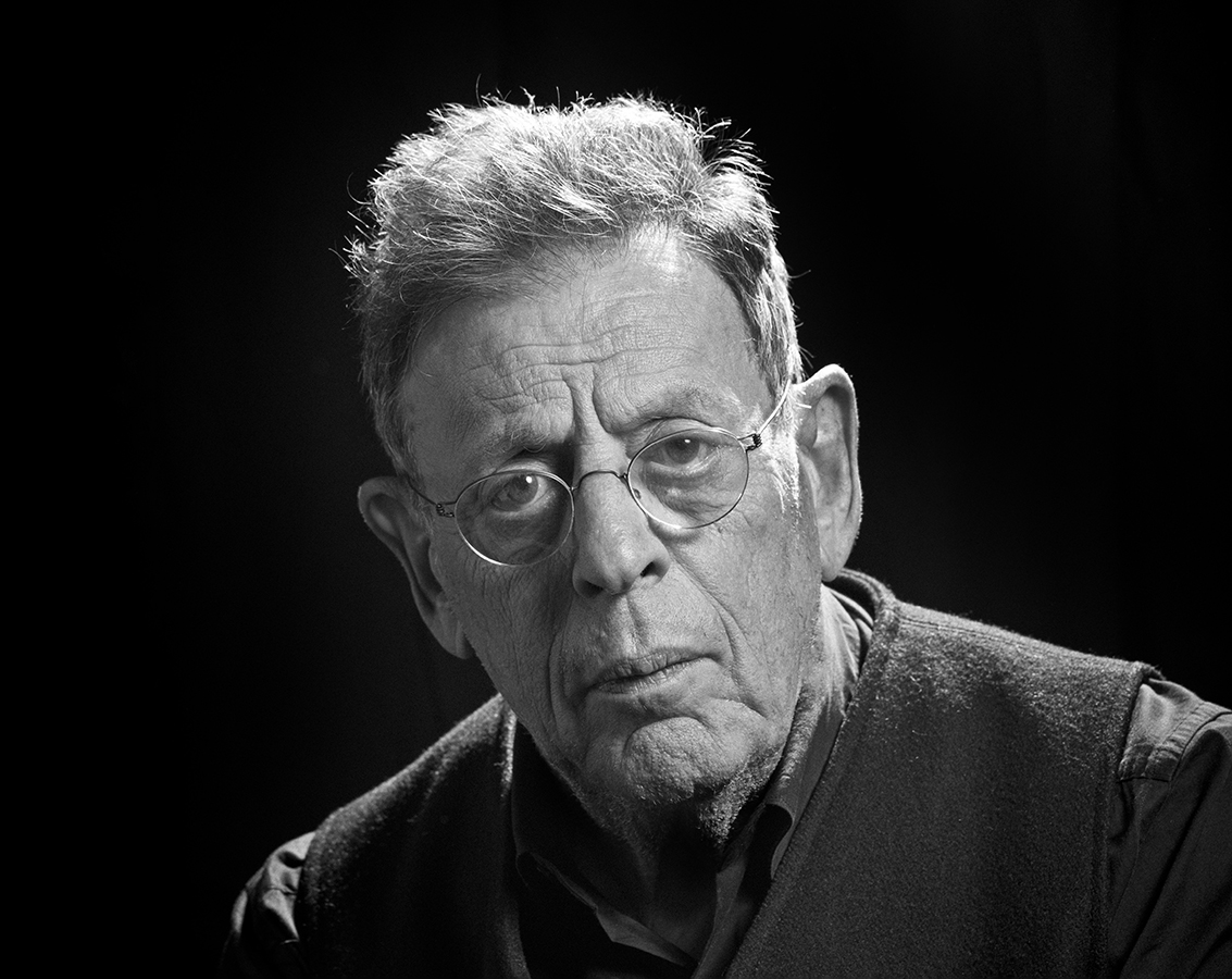 A black and white portrait of Philip Glass, an older man in a dark shirt and vest wearing glasses looking directly into the camera.