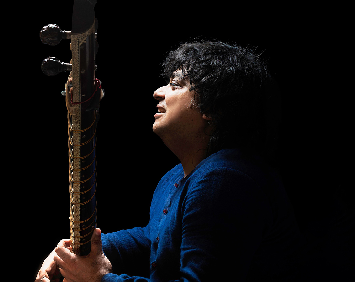 Sitarist Niladri Kumaar, a south Asian man dressed in blue sitting sideways and holding his instrument.