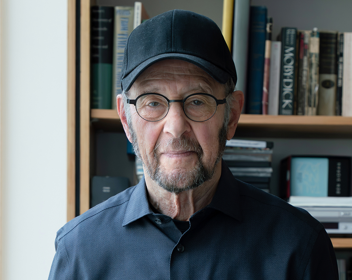 Composer Steve Reich, an older man with glasses and a baseball hat stands in front of a bookcase filled with books.