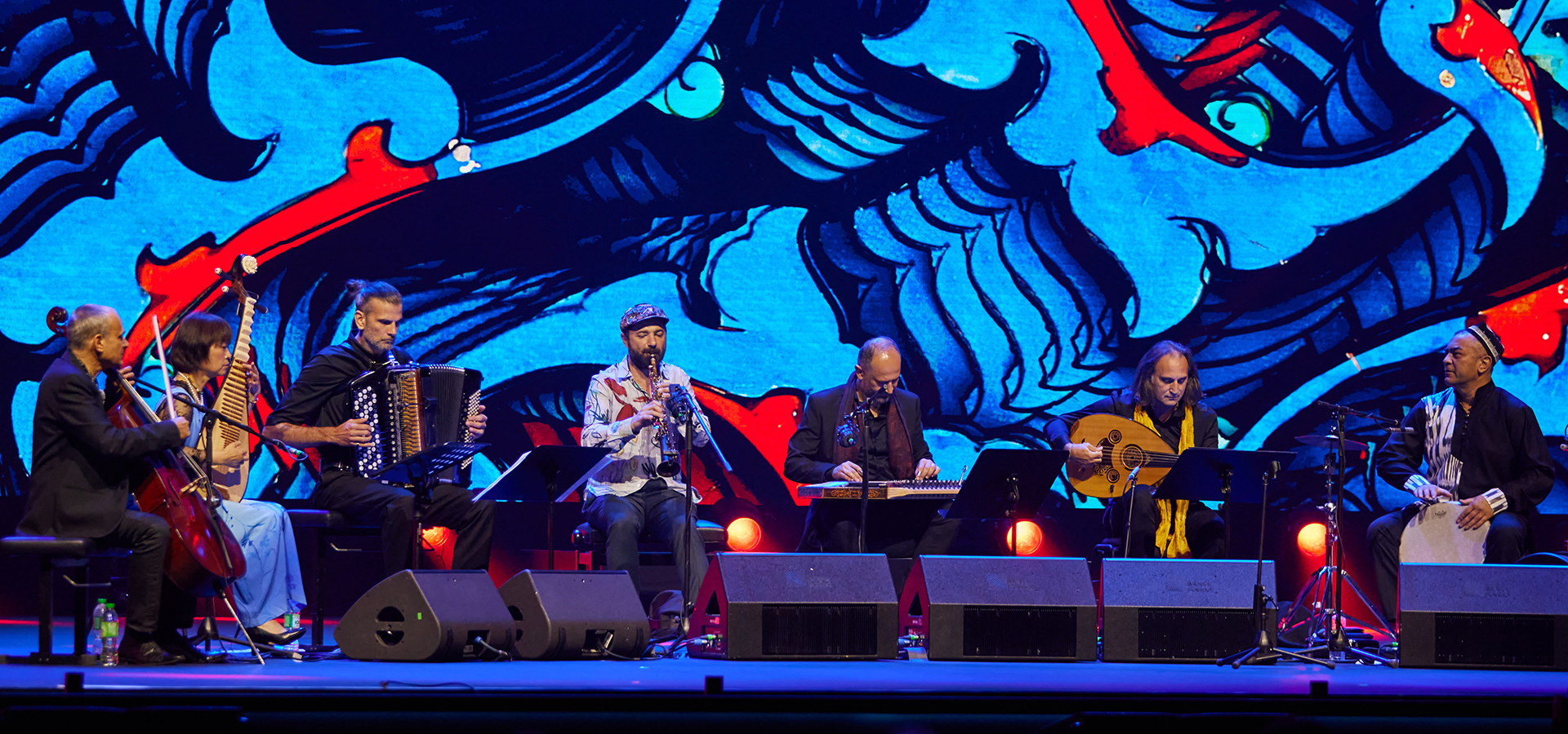 An ensemble photo of instrumentalists of Aga Khan Master Musicians in concert on stage in front of a blue and red backdrop.