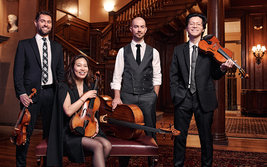 The four members of Dover Quartet -- three men and one women -- pose together with their instruments in a room with a wooden staircase.