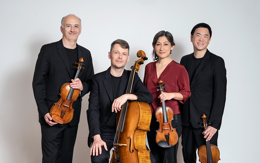 The four members of Takacs Quartet -- three men and one woman -- pose together with their instruments against a light gray background