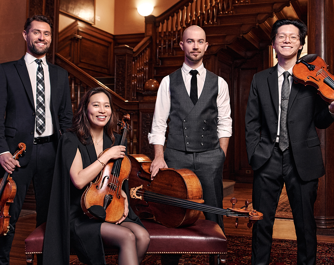 The four members of Dover Quartet -- three men and one women -- pose together with their instruments in a room with a wooden staircase.
