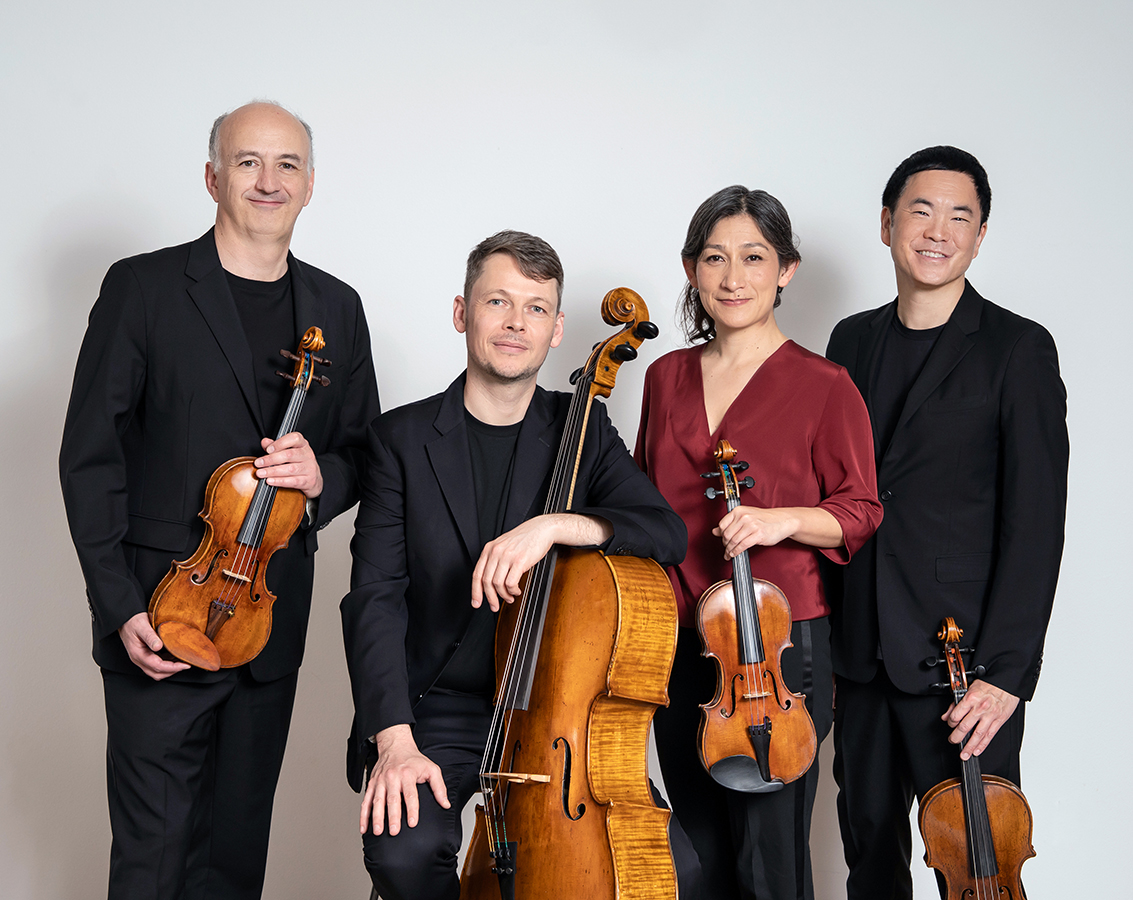 The four members of Takacs Quartet -- three men and one woman -- pose together with their instruments against a light gray background