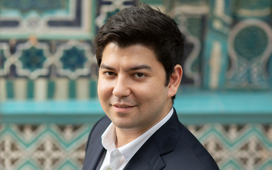 Pianist Behzod Abduraimov, an Uzbek man with dark hair wearing a dark suit jacket and white dress shirt, look into the camera against a blue and white tiled wall.