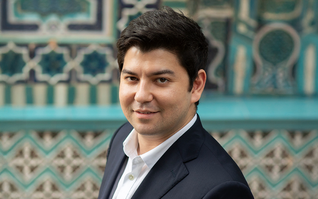 Pianist Behzod Abduraimov, an Uzbek man with dark hair wearing a dark suit jacket and white dress shirt, look into the camera against a blue and white tiled wall.