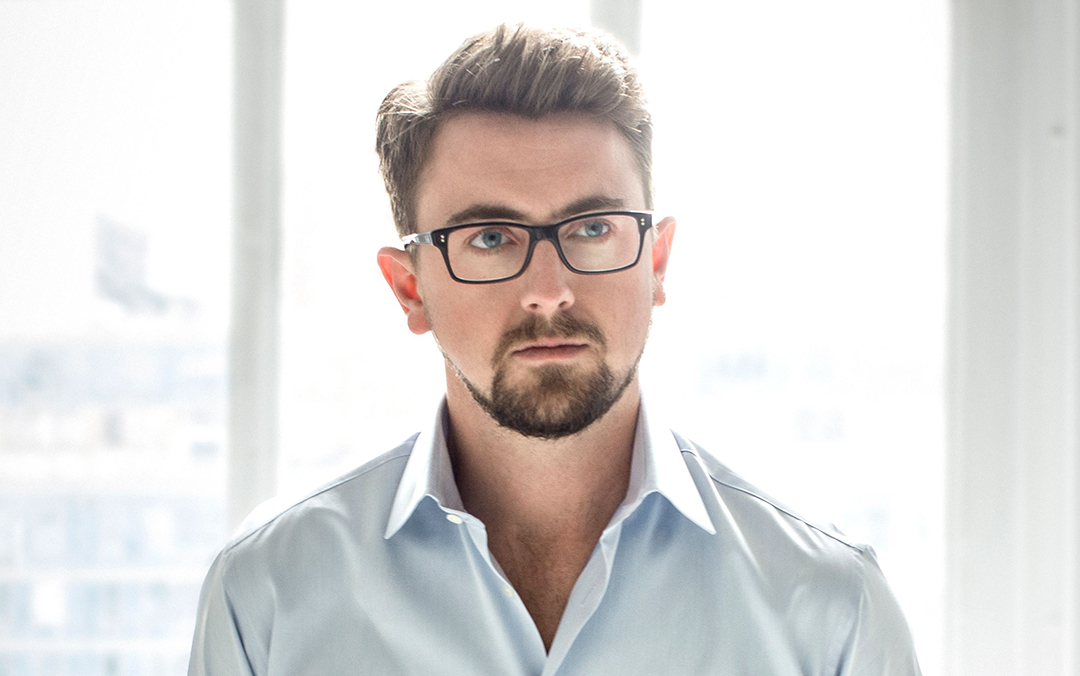 Tenor Ben Bliss, a white man wearing glasses and a light-colored shirt, stands in front of a window with natural light illuminating the scene.