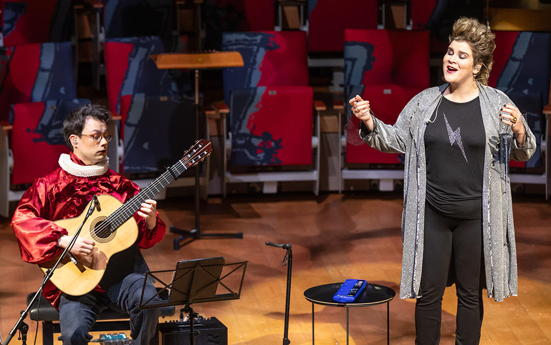 Mezzo-soprano Ema Nikolovska, a white woman wearing a long gray sweater over a black top adorned by a gray lightening bolt and black pants and boots, on singing stage with guitarist Sean Shibe, a mixed race man in glasses seated wearing a red top and Elizabethan white ruffled collar and jeans, playing a guitar.