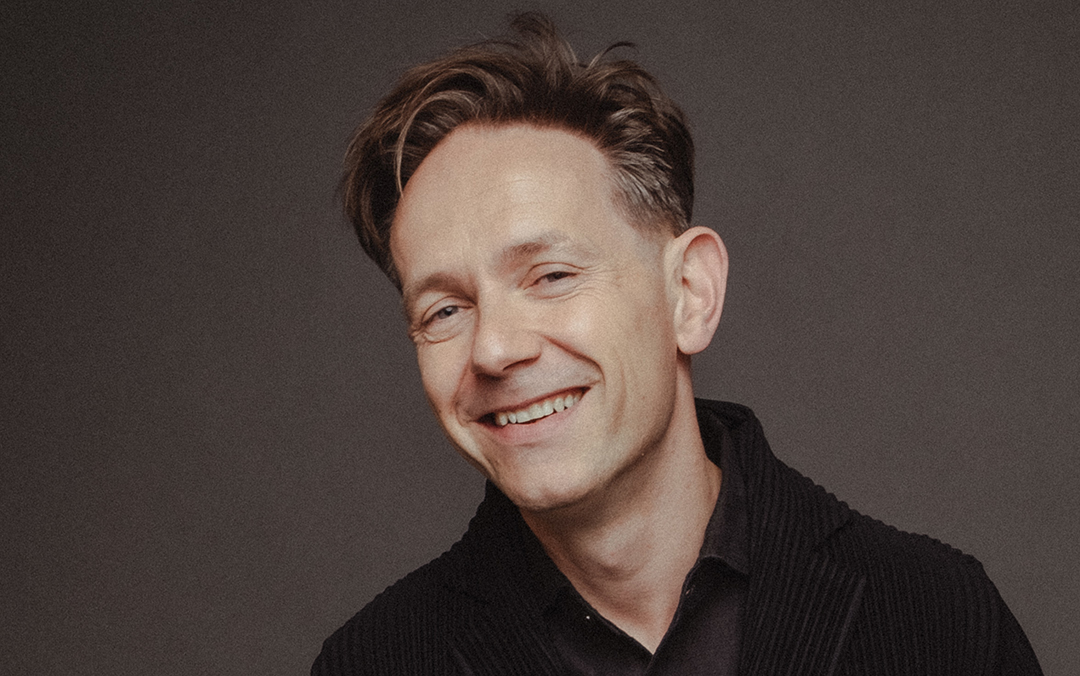 Countertenor Iestyn Davies, a smiling white man dressed in a black shirt and jacket, poses looks into the camera