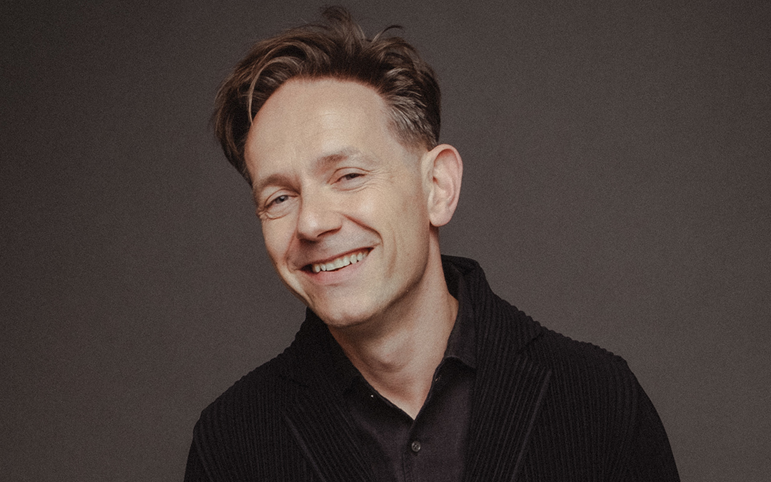 Countertenor Iestyn Davies, a smiling white man dressed in a black shirt and jacket, poses looks into the camera