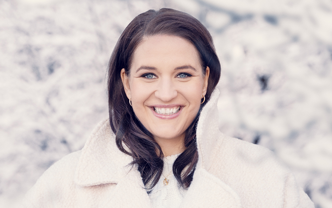 Soprano Lise Davidsen, a white woman with long brown hair dressed in a white sweater and white fleece coat, looks toward the camera against a snowy background