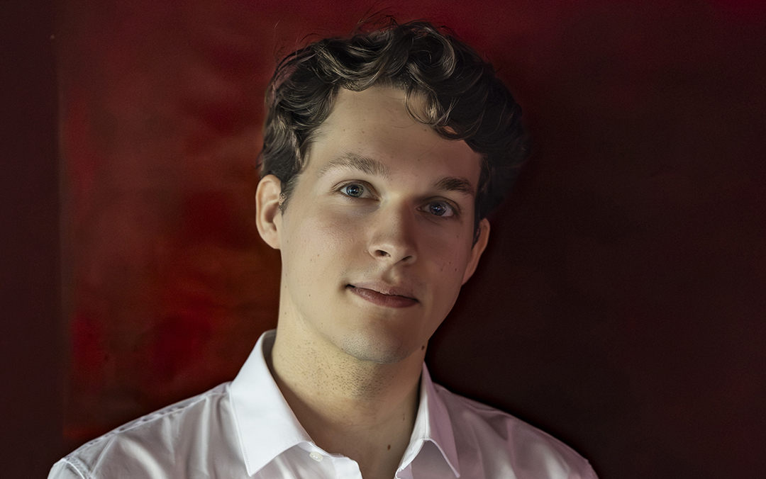 Pianist Lukas Sternath, a white young man in a white shirt against a dark red background, gazes directly at the camera with a slight smile.