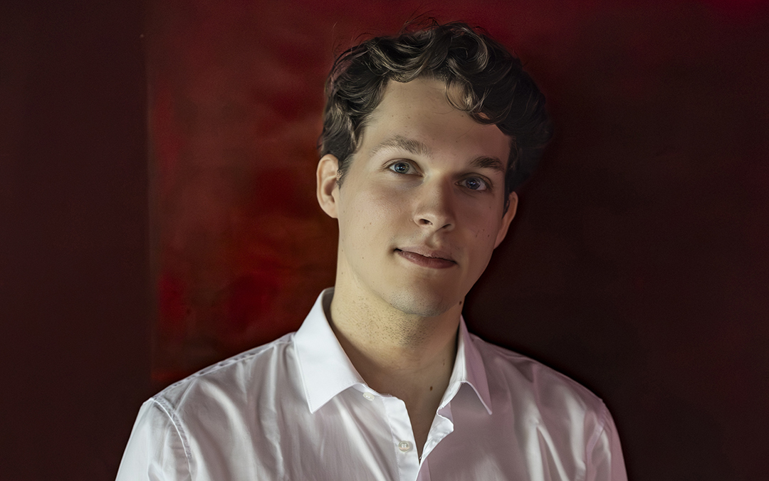 Pianist Lukas Sternath, a white young man in a white shirt against a dark red background, gazes directly at the camera with a slight smile.