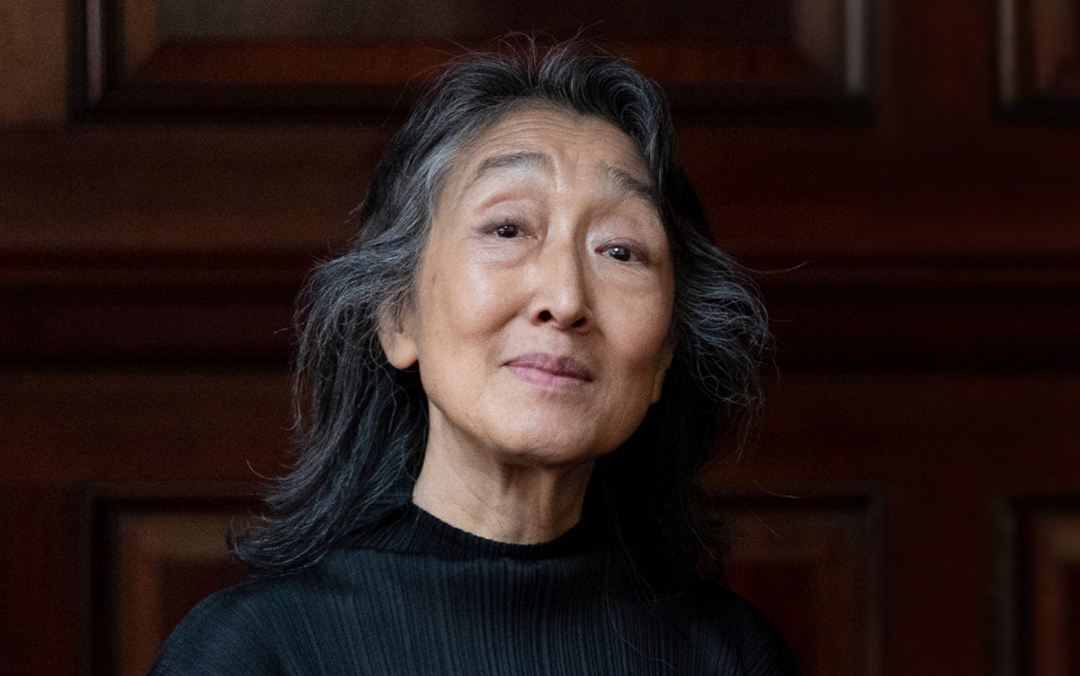 Pianist Mitsuko Uchida, an Asian woman with long silver and black hair wearing a black top, stands in front a dark wood background.