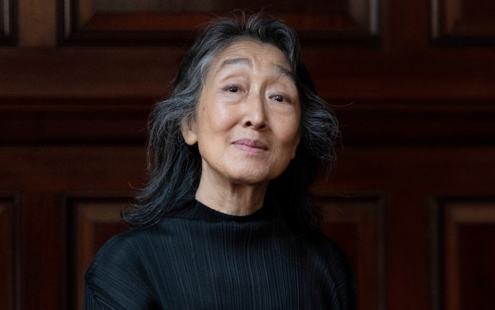 Pianist Mitsuko Uchida, an Asian woman with long silver and black hair wearing a black top, stands in front a dark wood background.