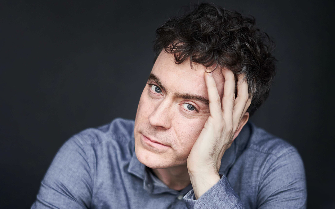 Pianist Paul Lewis, a white man with dark curly hair wearing a blue-gray shirt, leans forward with his head leaning on his hand against a gray background.