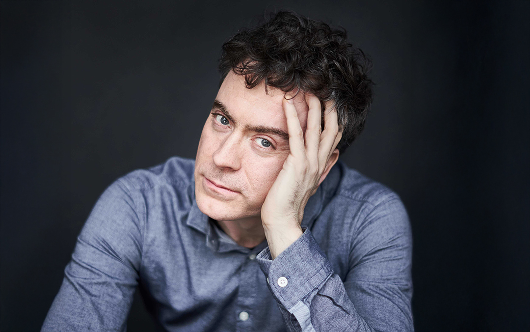 Pianist Paul Lewis, a white man with dark curly hair wearing a blue-gray shirt, leans forward with his head leaning on his hand against a gray background.