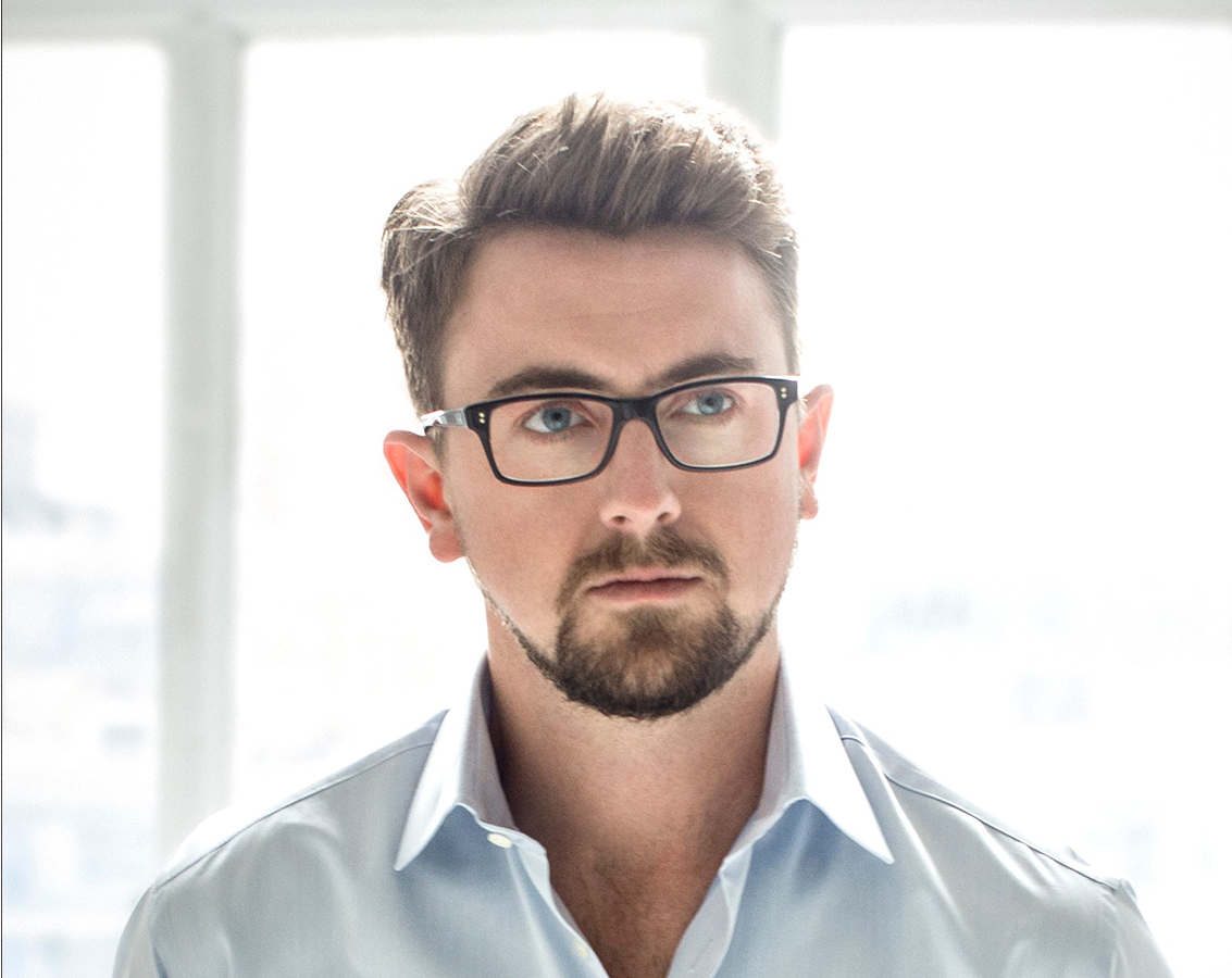 Tenor Ben Bliss, a white man wearing glasses and a light-colored shirt, stands in front of a window with natural light illuminating the scene.