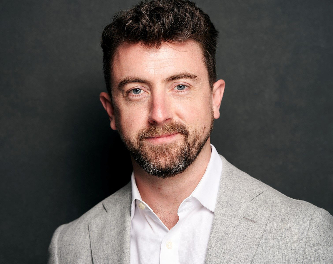 Photo of tenor Ben Bliss, a white man with wavy brown hair and slightly graying beard/moustache, in a gray suit jacket over a white button-down shirt against a dark gray background, looking into the camera