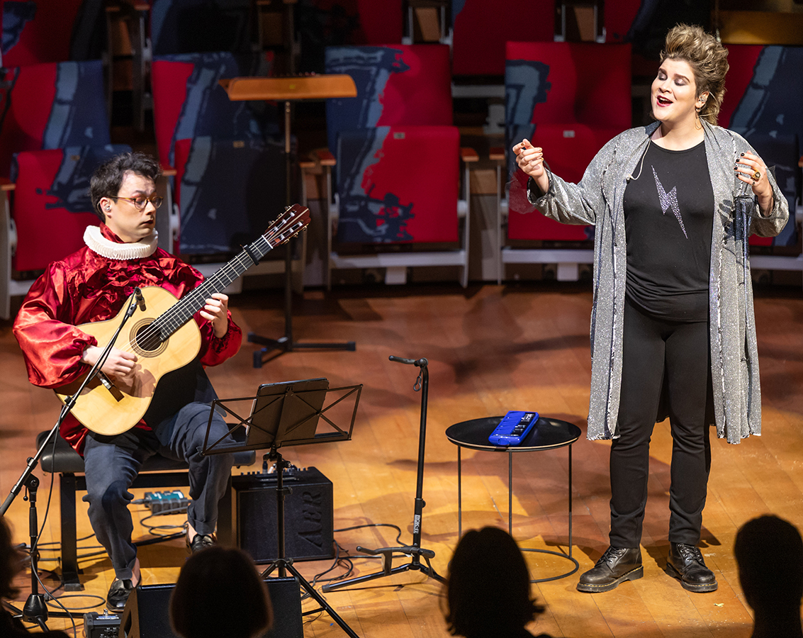 Mezzo-soprano Ema Nikolovska, a white woman wearing a long gray sweater over a black top adorned by a gray lightening bolt and black pants and boots, on singing stage with guitarist Sean Shibe, a mixed race man in glasses seated wearing a red top and Elizabethan white ruffled collar and jeans, playing a guitar.