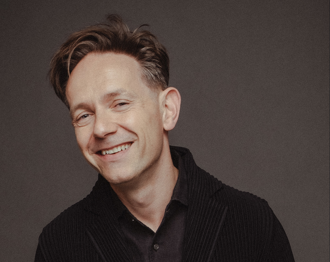 Countertenor Iestyn Davies, a smiling white man dressed in a black shirt and jacket, poses looks into the camera