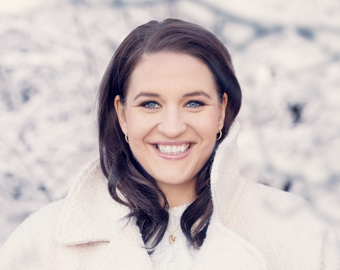 Soprano Lise Davidsen, a white woman with long brown hair dressed in a white sweater and white fleece coat, looks toward the camera against a snowy background
