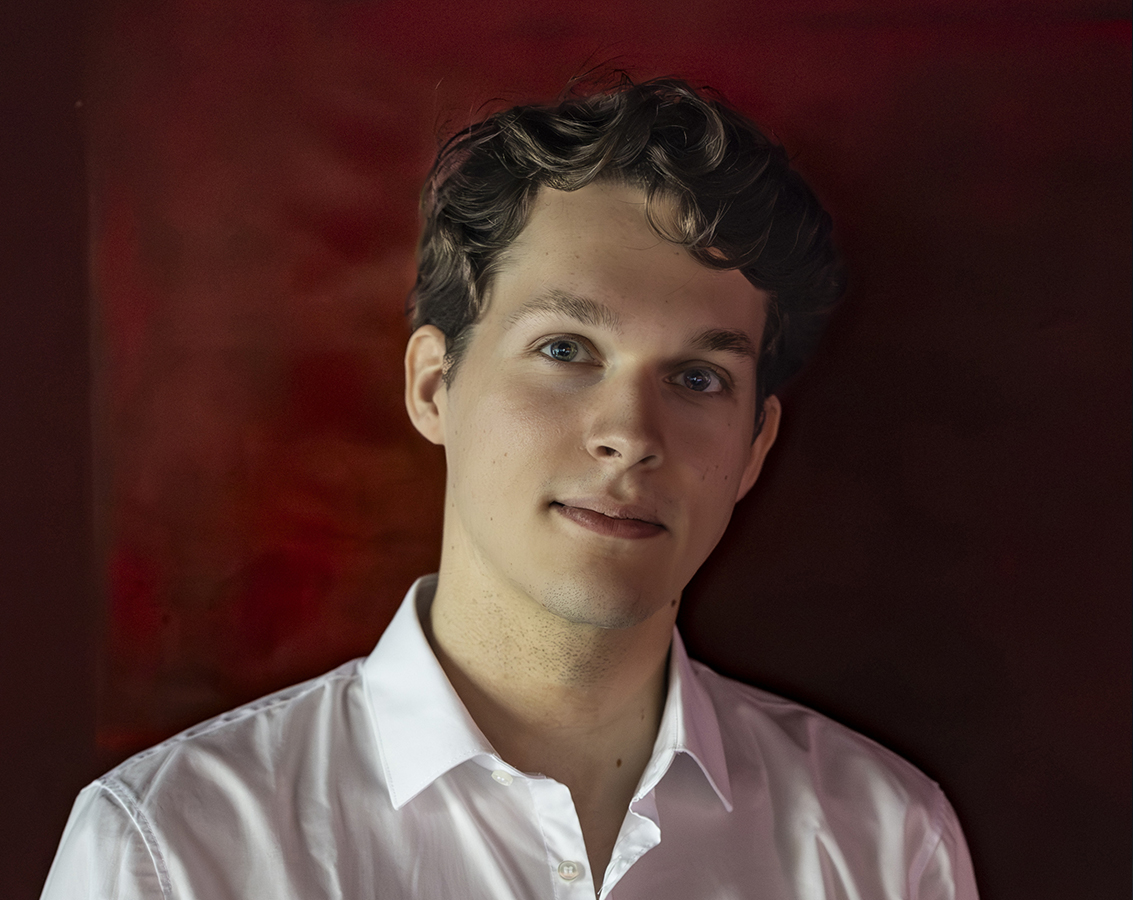 Pianist Lukas Sternath, a white young man in a white shirt against a dark red background, gazes directly at the camera with a slight smile.