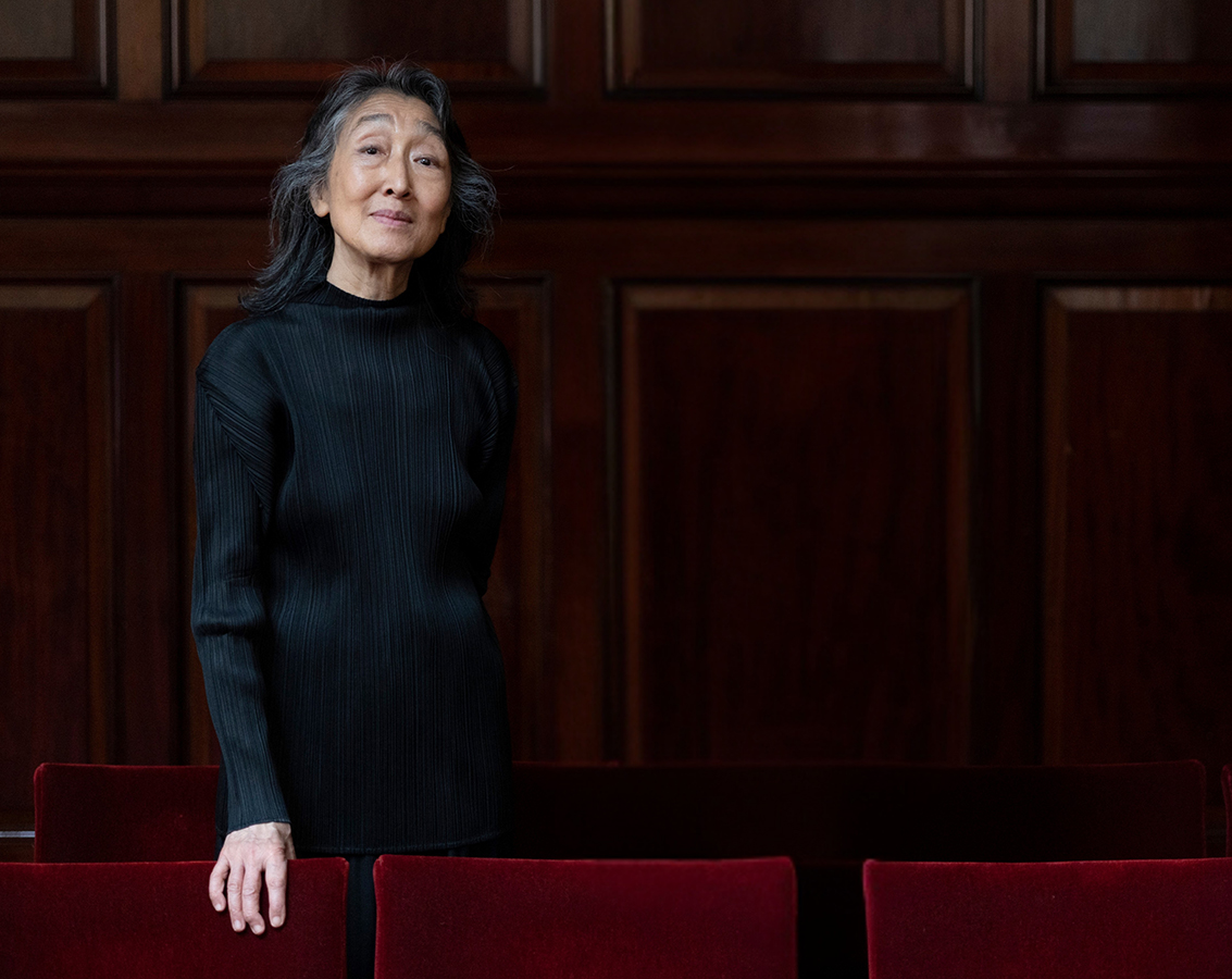 Pianist Mitsuko Uchida, an Asian woman with long silver and black hair wearing a black top, stands in front a dark wood background.