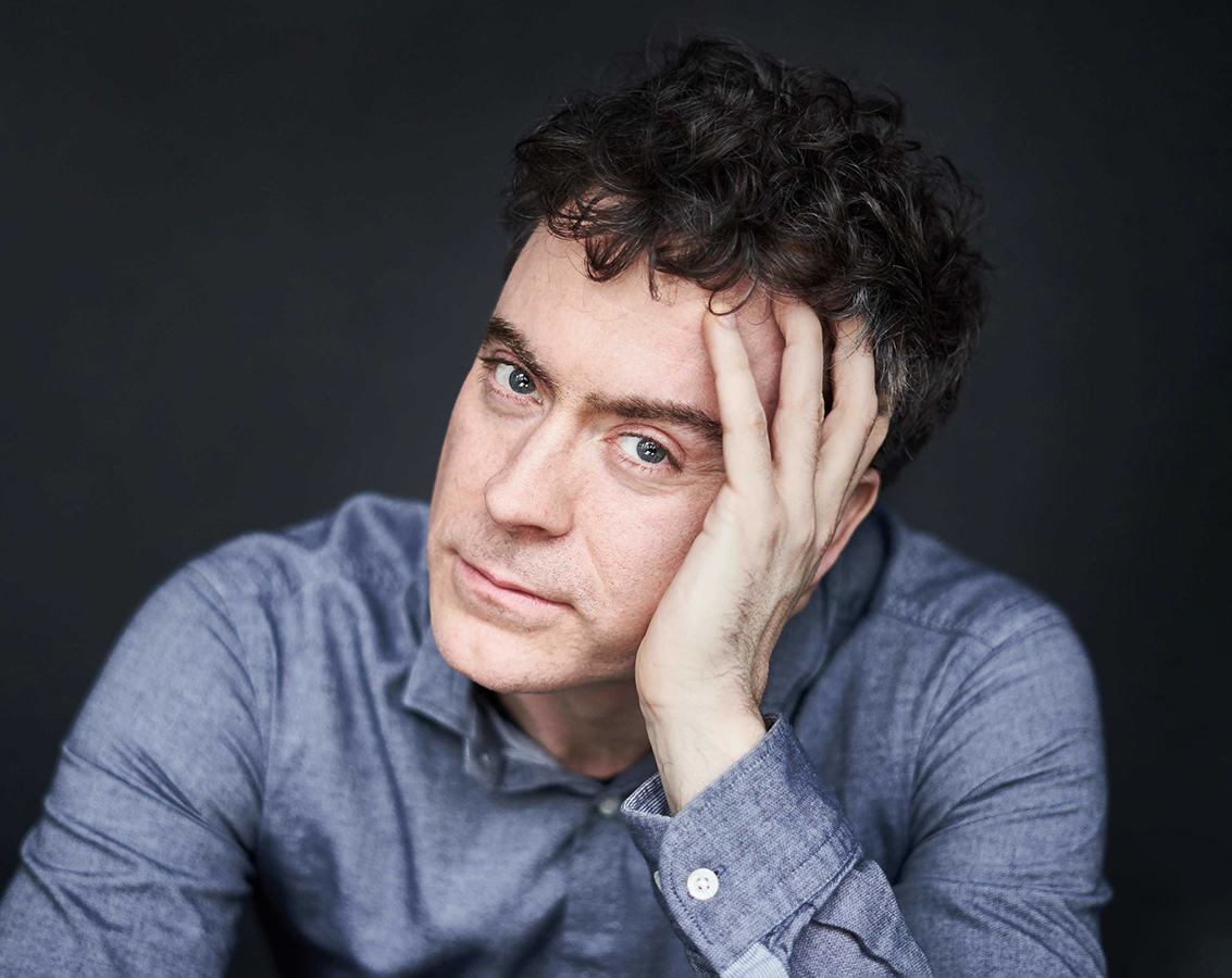 Pianist Paul Lewis, a white man with dark curly hair wearing a blue-gray shirt, leans forward with his head leaning on his hand against a gray background.