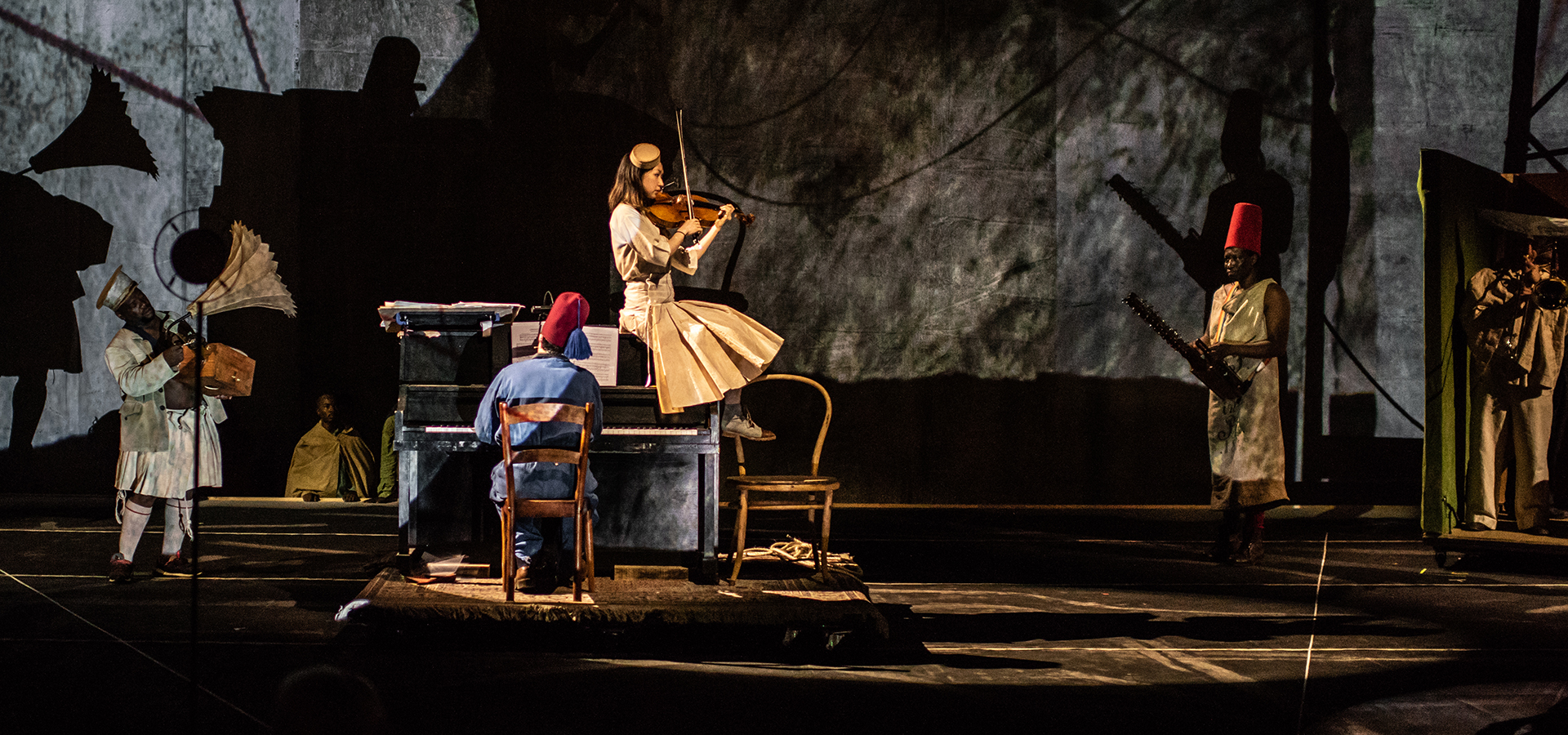A photo of a scene from William Kentridge's The Head and The Load, the actors playing several instruments against a multimedia backdrop.