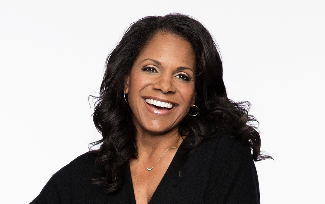 Award-winning vocalist and actor Audra McDonald, a black woman with long black hair, smiles while wearing a v-neck black top against a white background.