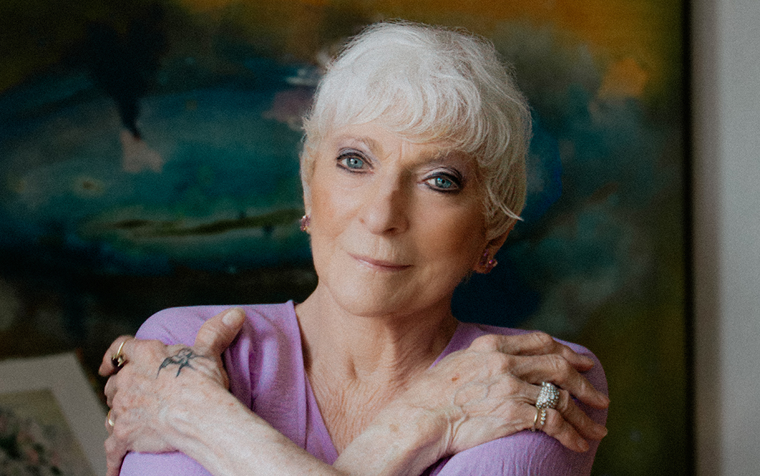 Folk singer Judy Collins, an older woman with white hair and visible hand tattoos wearing a light purple top against a colorful backdrop, looks into the camera.
