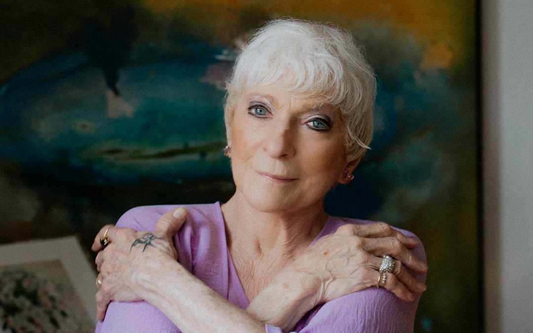 Folk singer Judy Collins, an older woman with white hair and visible hand tattoos wearing a light purple top against a colorful backdrop, looks into the camera.