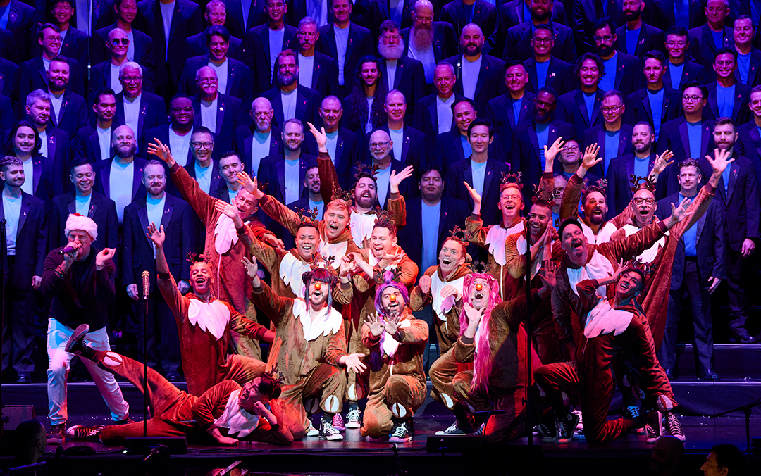 A lively performance photo of San Francisco Gay Men's Chorus, a small group of performers centered in reindeer costumes in front with the large chorus singing behind them.
