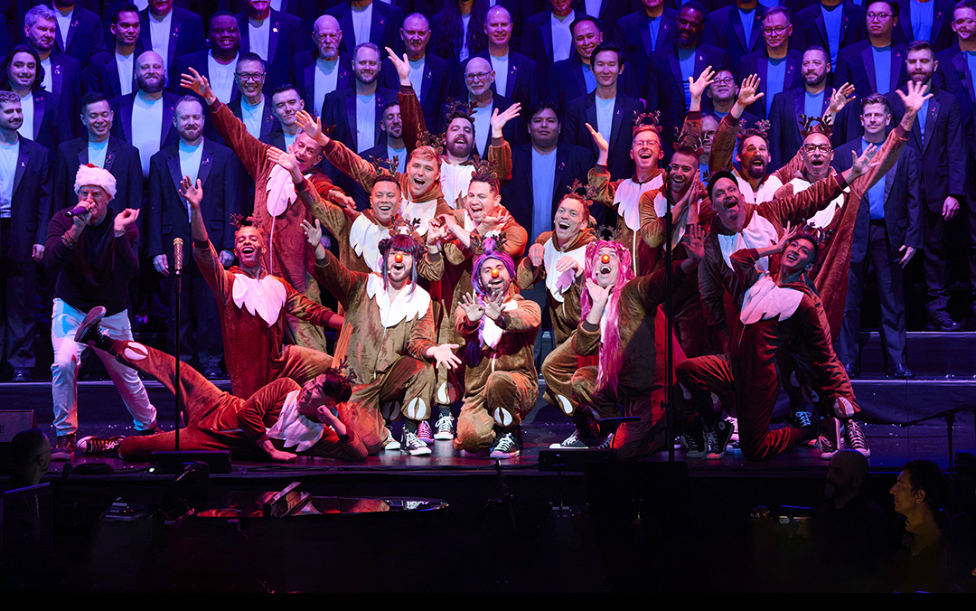 A lively performance photo of San Francisco Gay Men's Chorus, a small group of performers centered in reindeer costumes in front with the large chorus singing behind them.