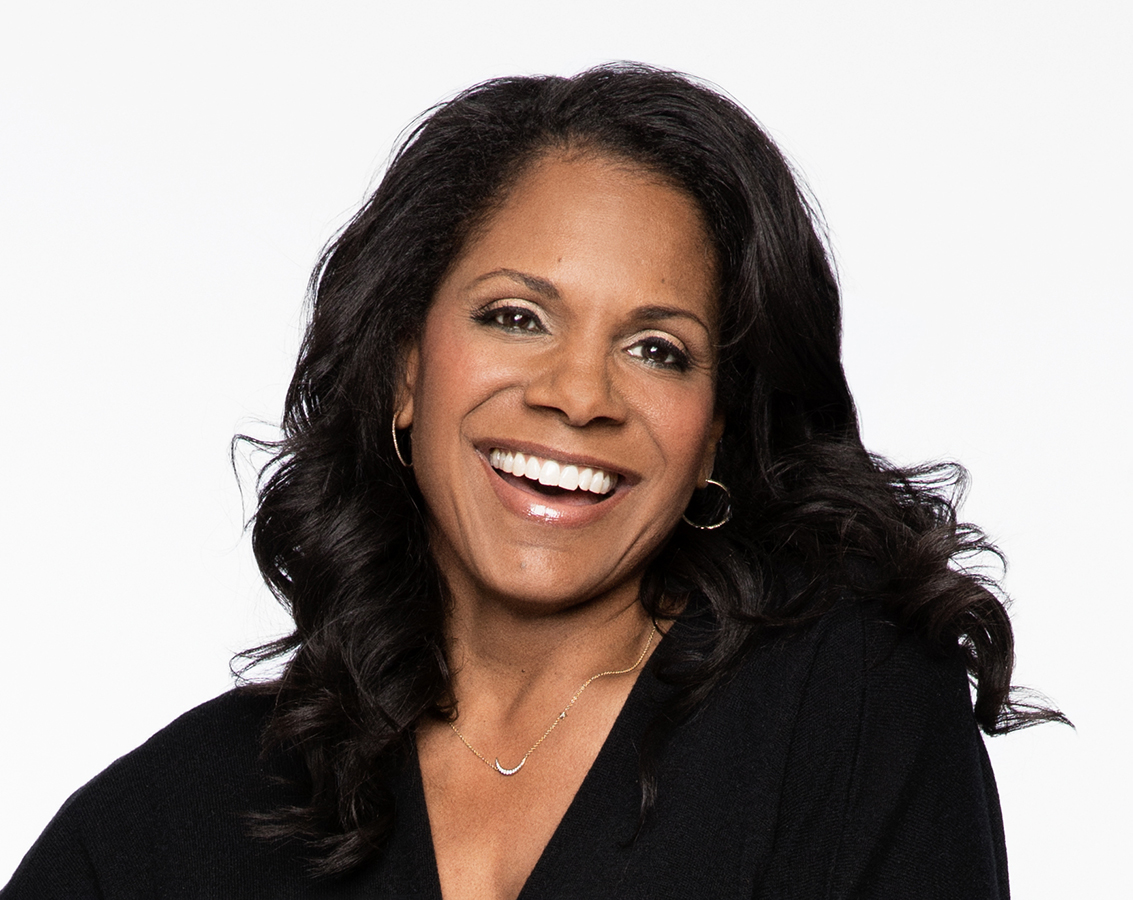 Award-winning vocalist and actor Audra McDonald, a black woman with long black hair, smiles while wearing a v-neck black top against a white background.