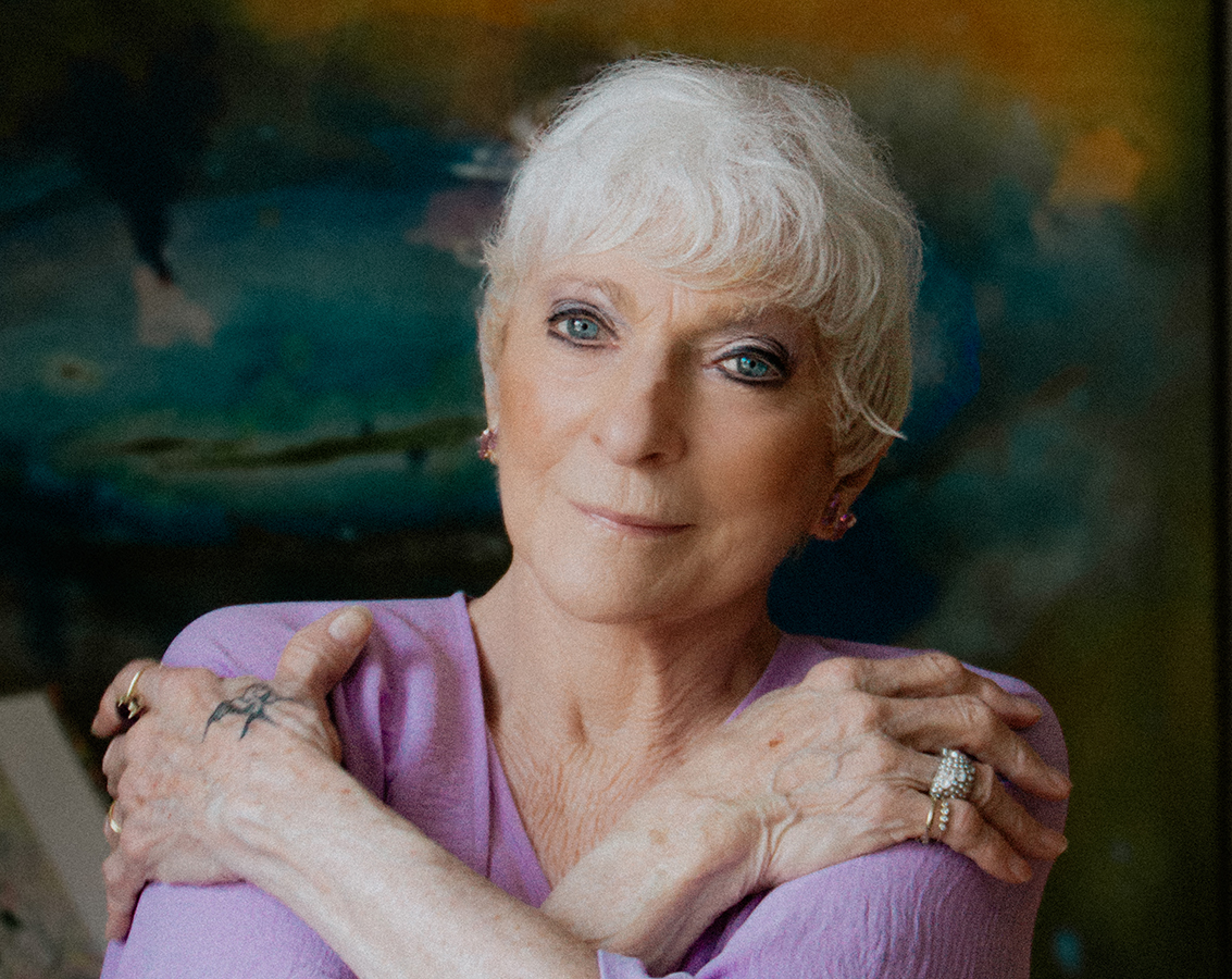 Folk singer Judy Collins, an older woman with white hair and visible hand tattoos wearing a light purple top against a colorful backdrop, looks into the camera.