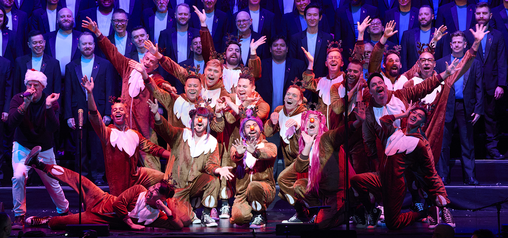A lively performance photo of San Francisco Gay Men's Chorus, a small group of performers centered in reindeer costumes in front with the large chorus singing behind them.