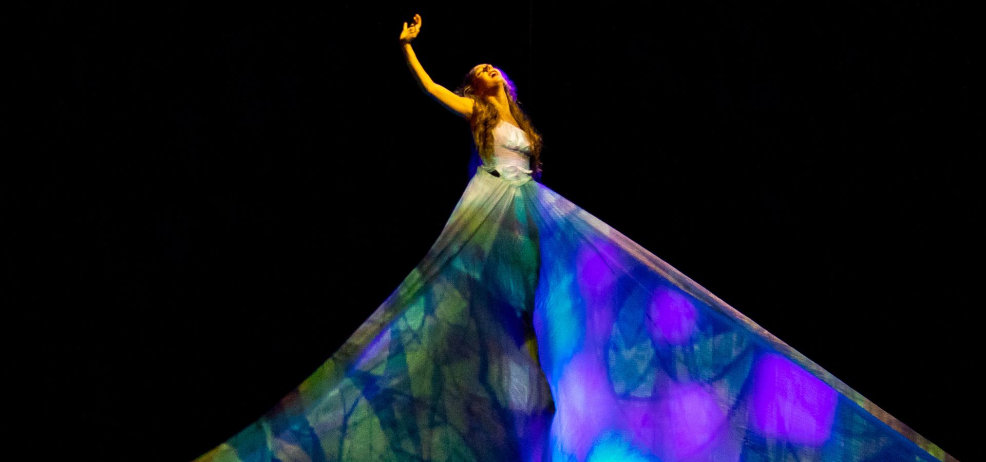 A member of MOMIX reaching up high in the air, wearing a large dress with a colorful stained glass-like pattern.