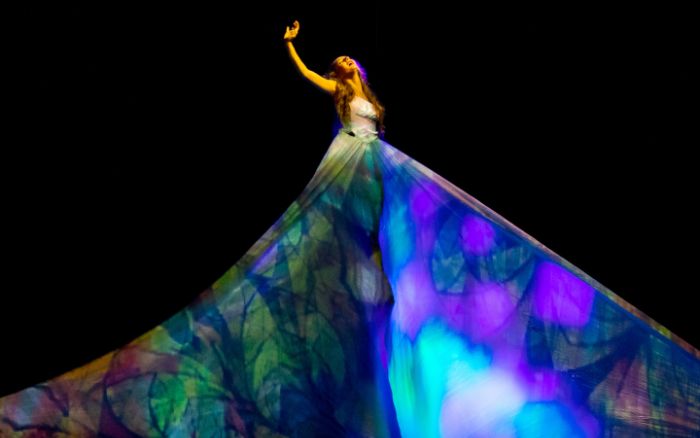 A member of MOMIX reaching up high in the air, wearing a large dress with a colorful stained glass-like pattern.