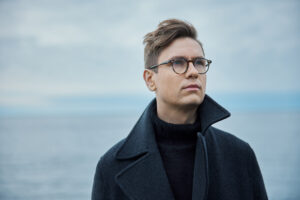 Pianist Vikingur Olafsson wears a collared coat and glasses in this close up photo, with a blue sky behind him.